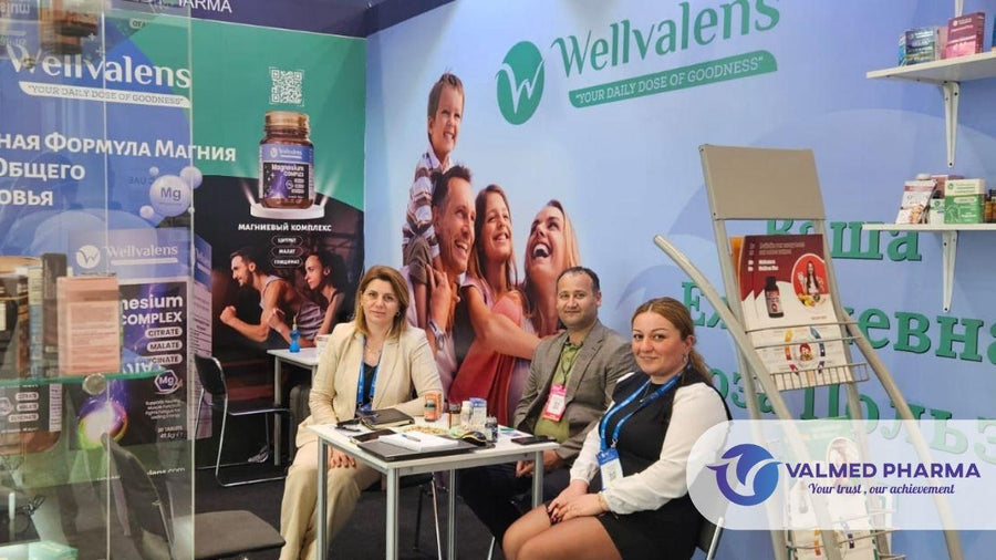 Three people seated at a Wellvalens exhibition booth with supplement displays, brochures and Valmed Pharma branding.