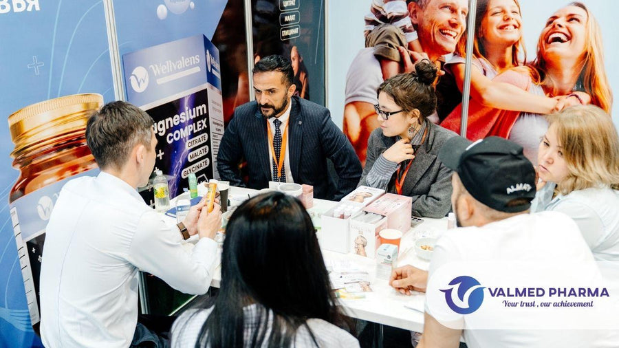 Visitors meeting around a table with supplement samples at a Wellvalens exhibition booth, with Valmed Pharma branding.