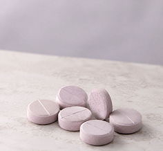 Six round, light pinkish-purple tablets with score lines, arranged on a light gray textured surface against a soft gray background.