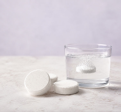 Three white round tablets are arranged beside a clear glass of water on a light stone surface, with one tablet dissolving in the liquid against a soft pale lavender background.