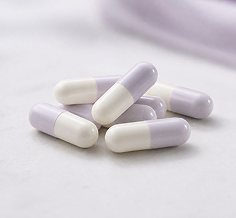 Several lavender and white capsule pills arranged on a soft white surface with a blurred lavender fabric in the background.