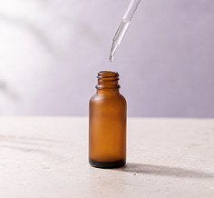 An empty amber glass dropper bottle with a glass pipette hovering above it, set on a light beige surface against a soft, out-of-focus lavender background.