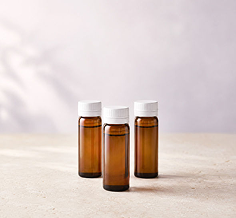 Three small amber glass bottles with white caps arranged on a light beige surface against a soft, pale gray background.