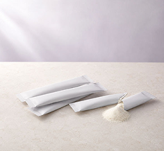 Four white sachets of powder are arranged on a light beige surface with a small pile of the white powder in front of them. The background is a soft, light lavender-gray gradient.
