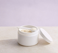 A white cosmetic jar with its lid off, showing a light beige cream or powder inside, with a small metal spoon resting in the container. The jar is placed on a light beige surface against a soft lavender background.