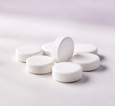 Six white round tablets or pills arranged on a soft light gray to white surface, with one pill standing upright in the center.